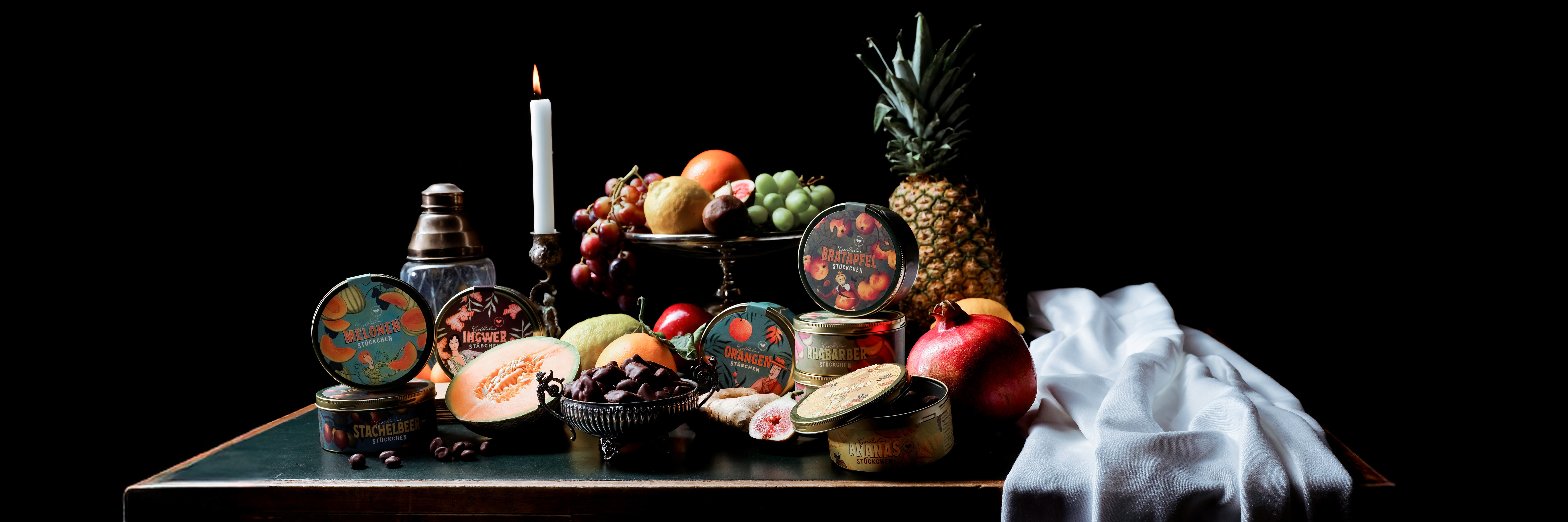 Kandierte Schokoladenfrüchte auf einem Tisch mit frischem Obst, einer Kerze und einem weißem Tuch, welches hübsch drapiert wurde.