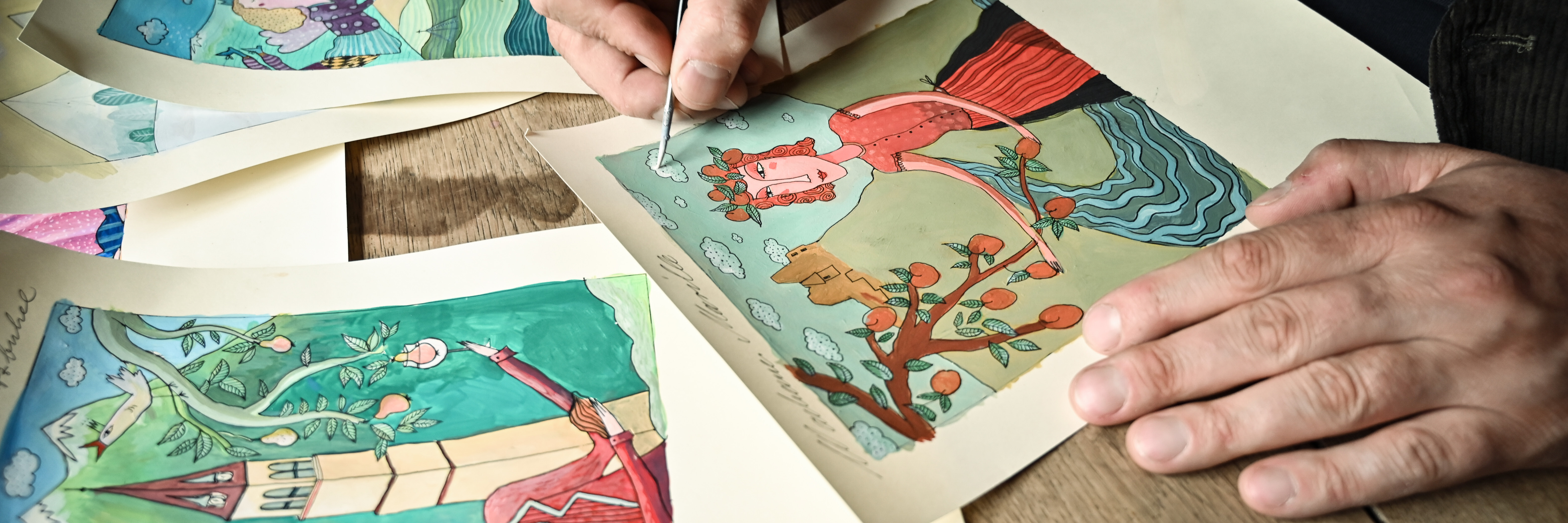 Verschiedene Illustrationen  liegen auf einem Tisch. Alex Kühn vollendet mit Farbe und Pinsel eine Illustration.