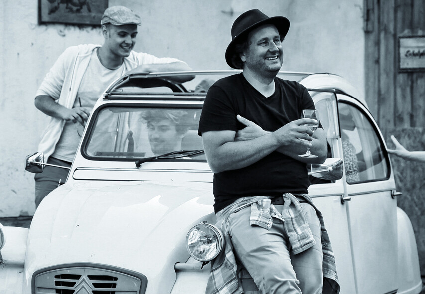 Alex Kühn lehnt gegen Auto, eine alte Ente, und hält ein Glas Wein in der Hand. Im Hintergrund stehen Gäste.
