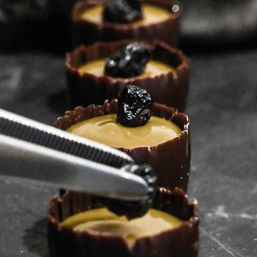 Goldhelm Pralinen liegen auf einer Marmorplatte und jemand veredelt mit einer Pinzette diese mit Trockenobst.